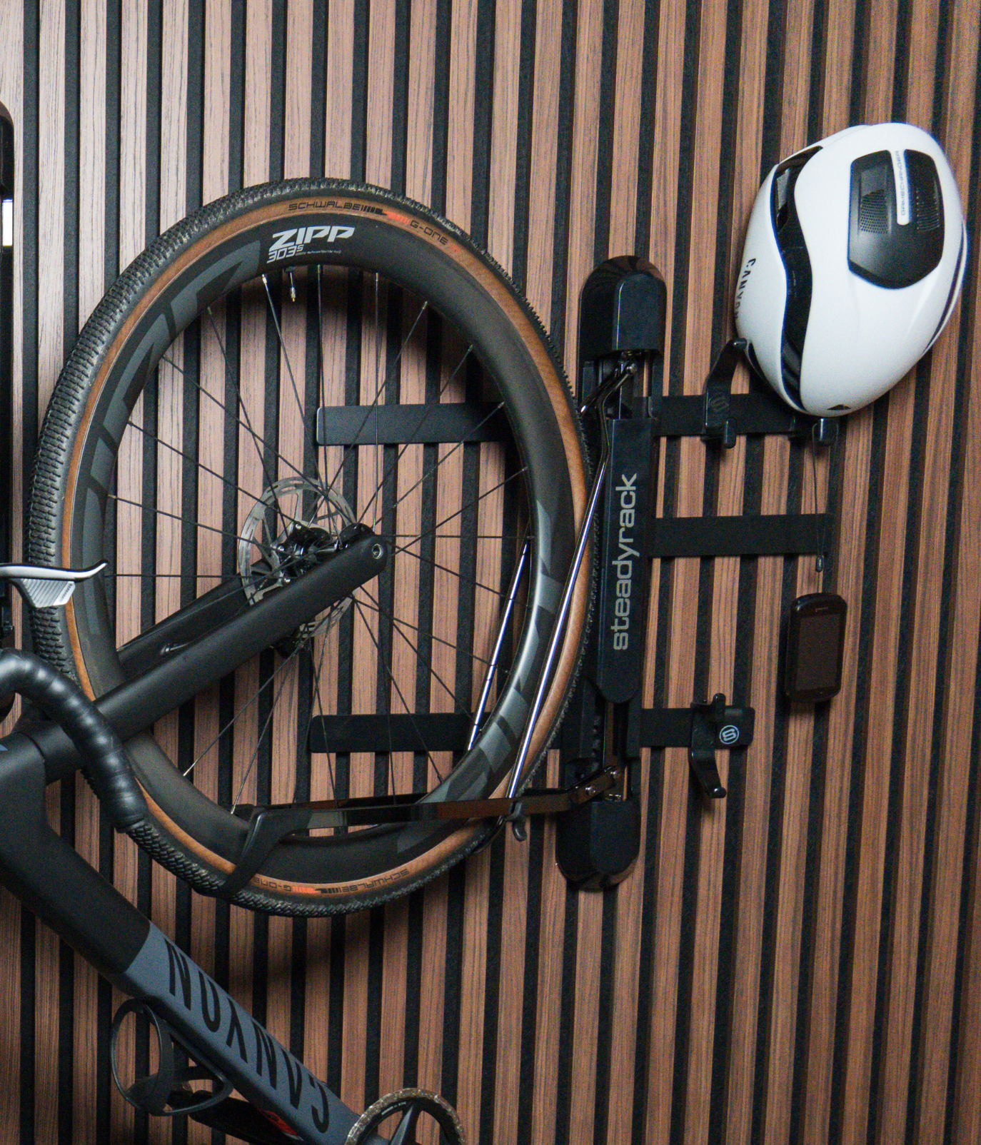 A Steadyrack bike rack on a wooden slat wall with a road bike stored. Attached to the rack is black metal brackets and a hook storing a helmet. 